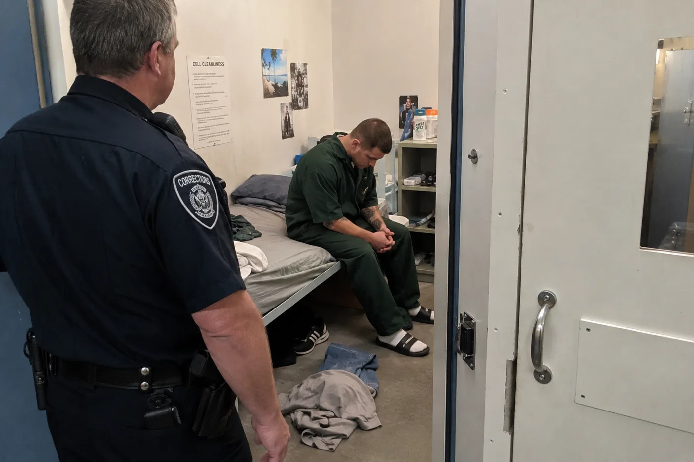 A correctional officer stands at the open door of a cell. Inside, a man in green prison clothing sits on the edge of an unmade bed, looking down. Clothes lie on the floor.